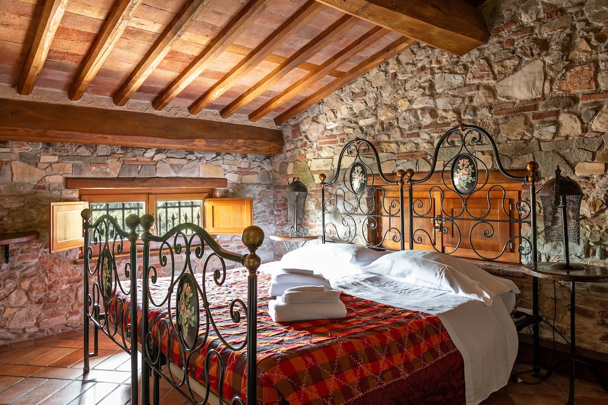 A bedroom with a stone wall, a wooden ceiling, a metal bed frame, and a red plaid bedspread.