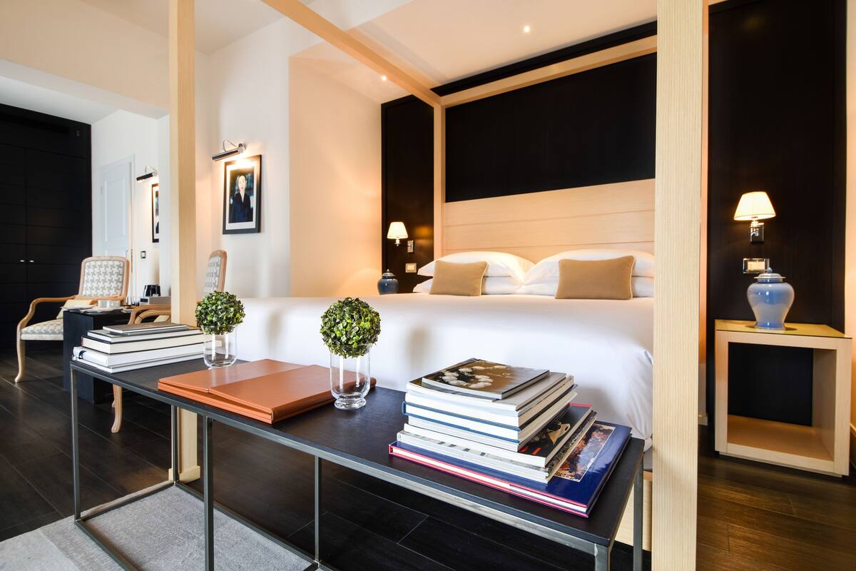 A modern hotel room with a large bed, bedside tables, a desk with books, and a small plant.