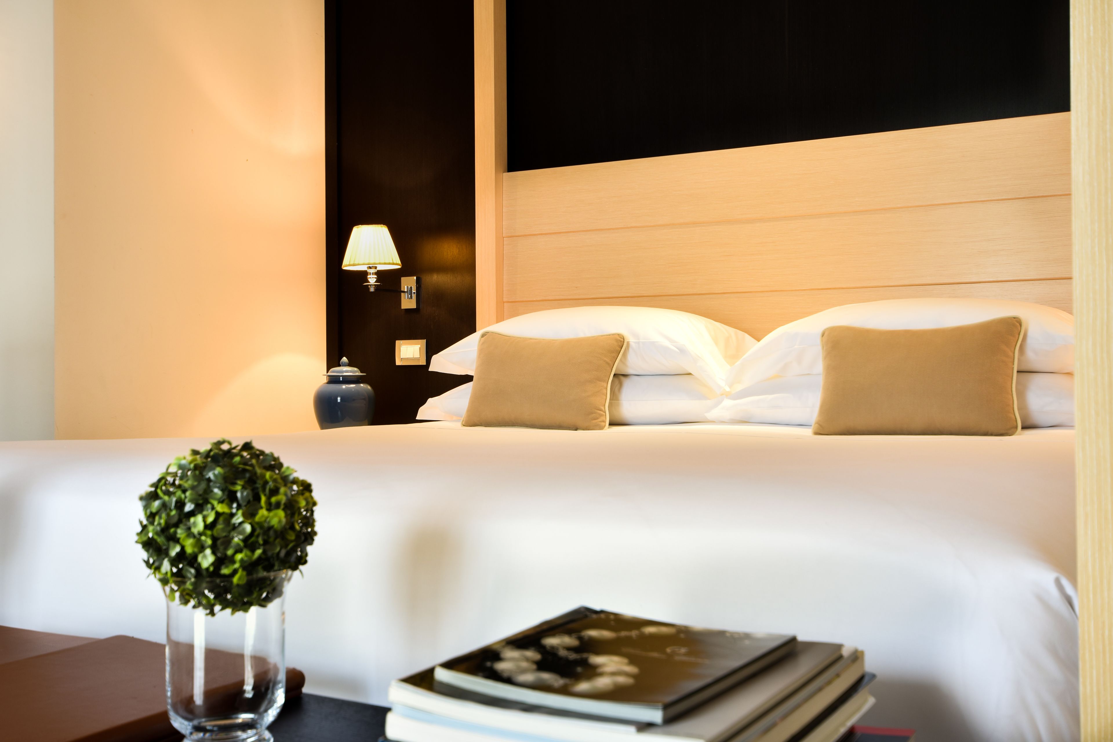 A neatly made bed with white and beige pillows, a bedside table with a lamp, and a small potted plant.