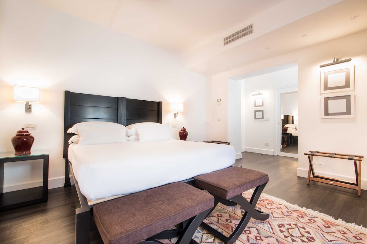 A modern hotel room with a large bed, bedside tables, a bench, and a patterned rug.