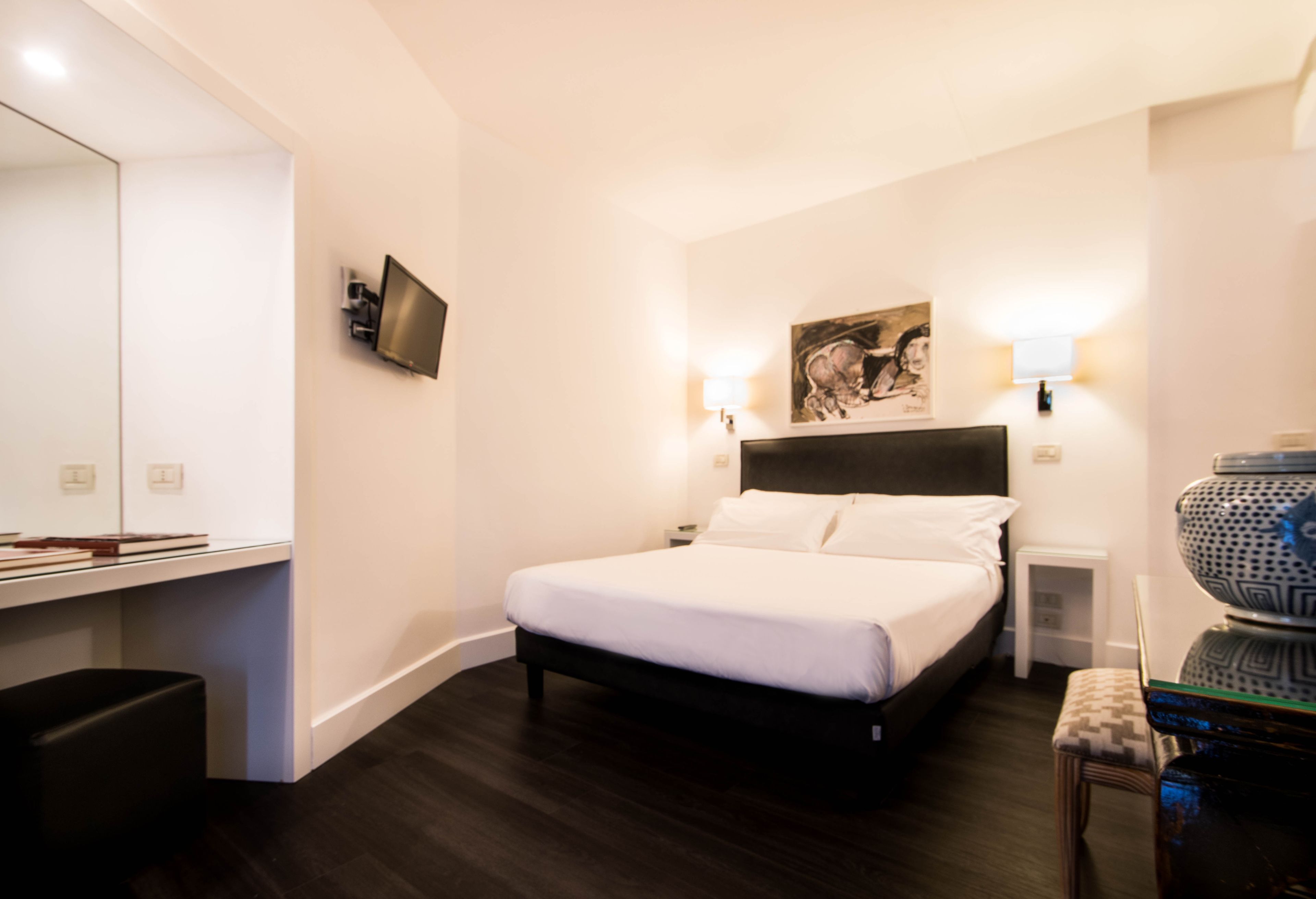 A modern hotel room with a large bed, bedside lamps, a flat-screen TV, a painting on the wall, and a small table with a vase.