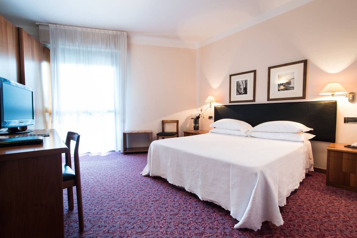 A hotel room with a bed, a desk with a computer, a chair, a lamp, and two framed pictures on the wall.