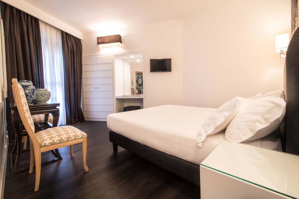 A hotel room with a bed, a chair, a desk, a mirror, and a TV.