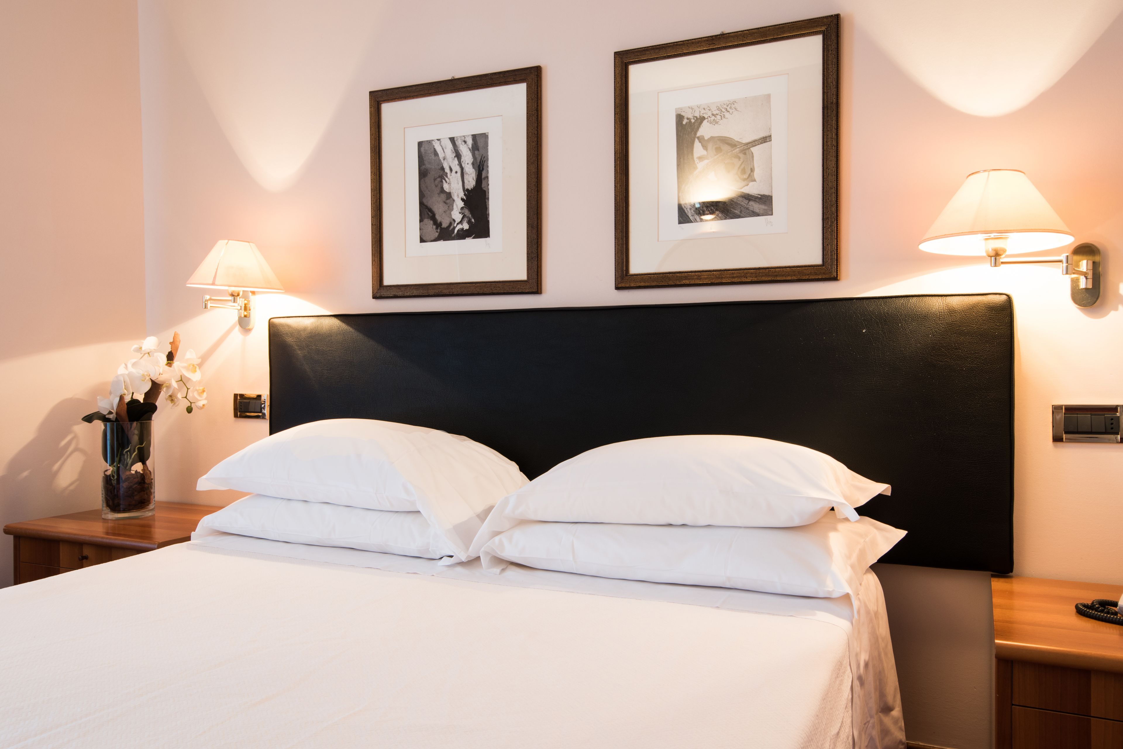 A neatly made bed with white linens, two framed pictures on the wall, and two bedside lamps.