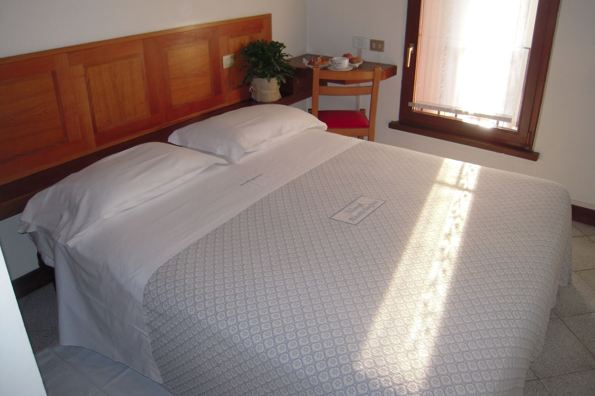 A neatly made bed with a patterned bedspread, a wooden headboard, a small table with a plant, and a window with curtains.