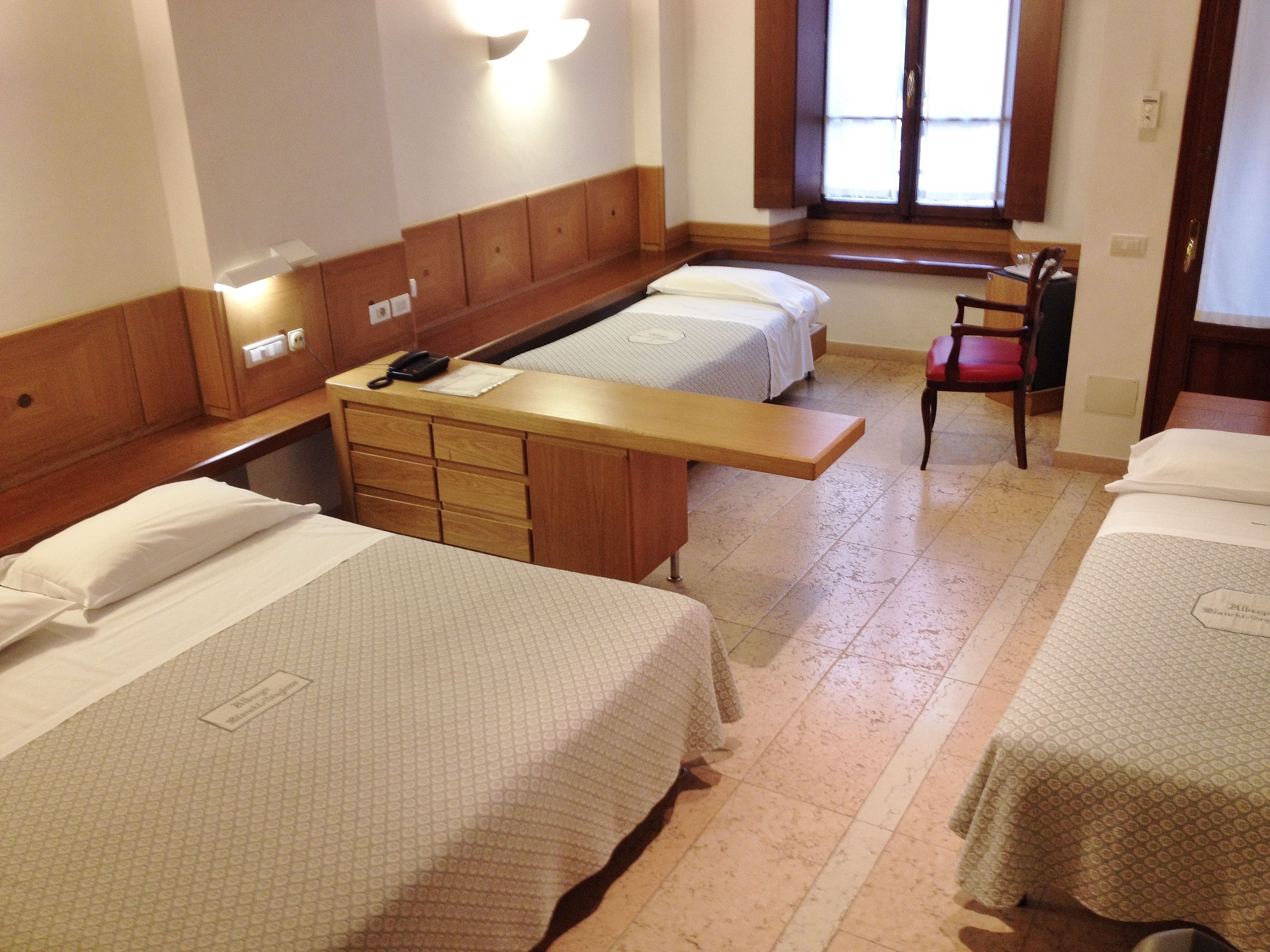 Habitación de hotel con tres camas, un escritorio de madera, una silla y un ventanal con persianas.
