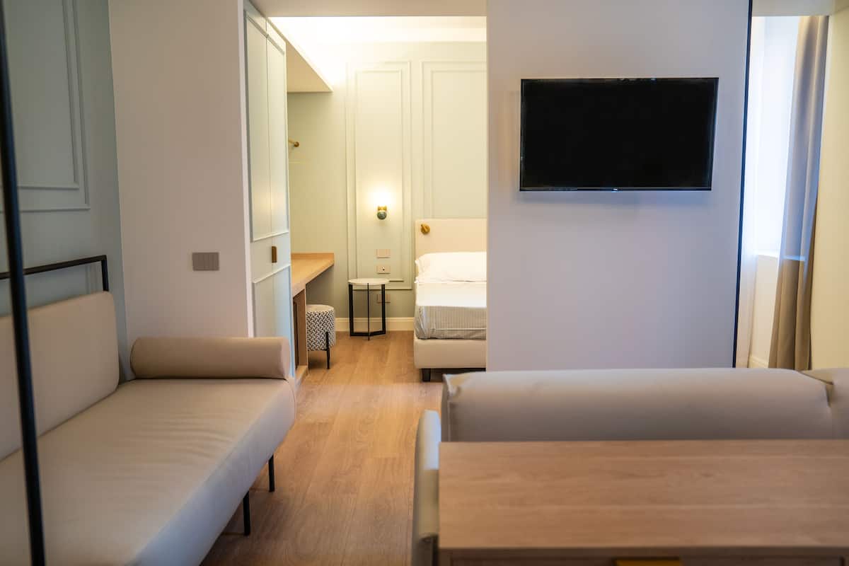 A compact hotel room with two beds, a small table, a TV, and a bathroom with a mirror and light fixture.