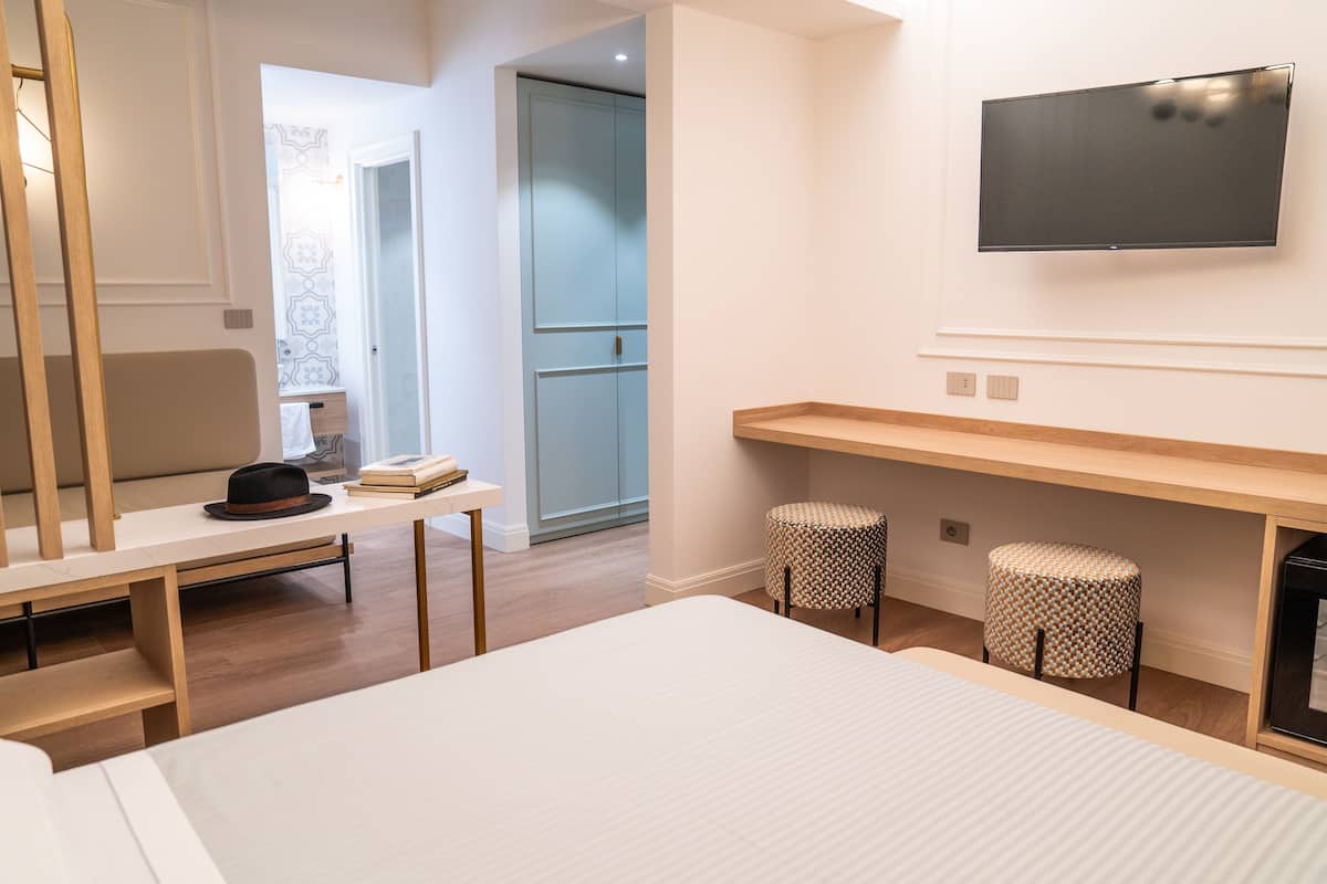 A modern hotel room with a bed, a desk, a TV mounted on the wall, and a small stool.