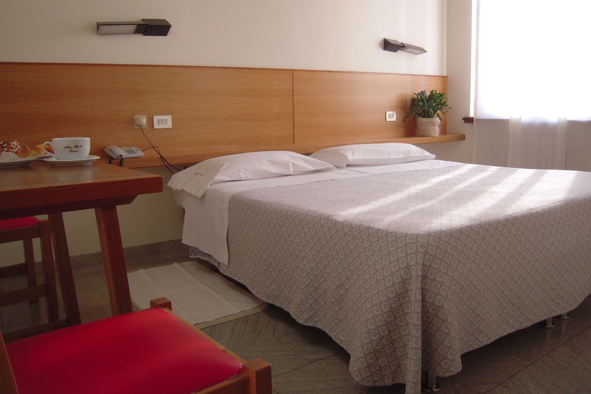 A hotel room with a bed, a desk with a phone, a cup of coffee, and a plant.