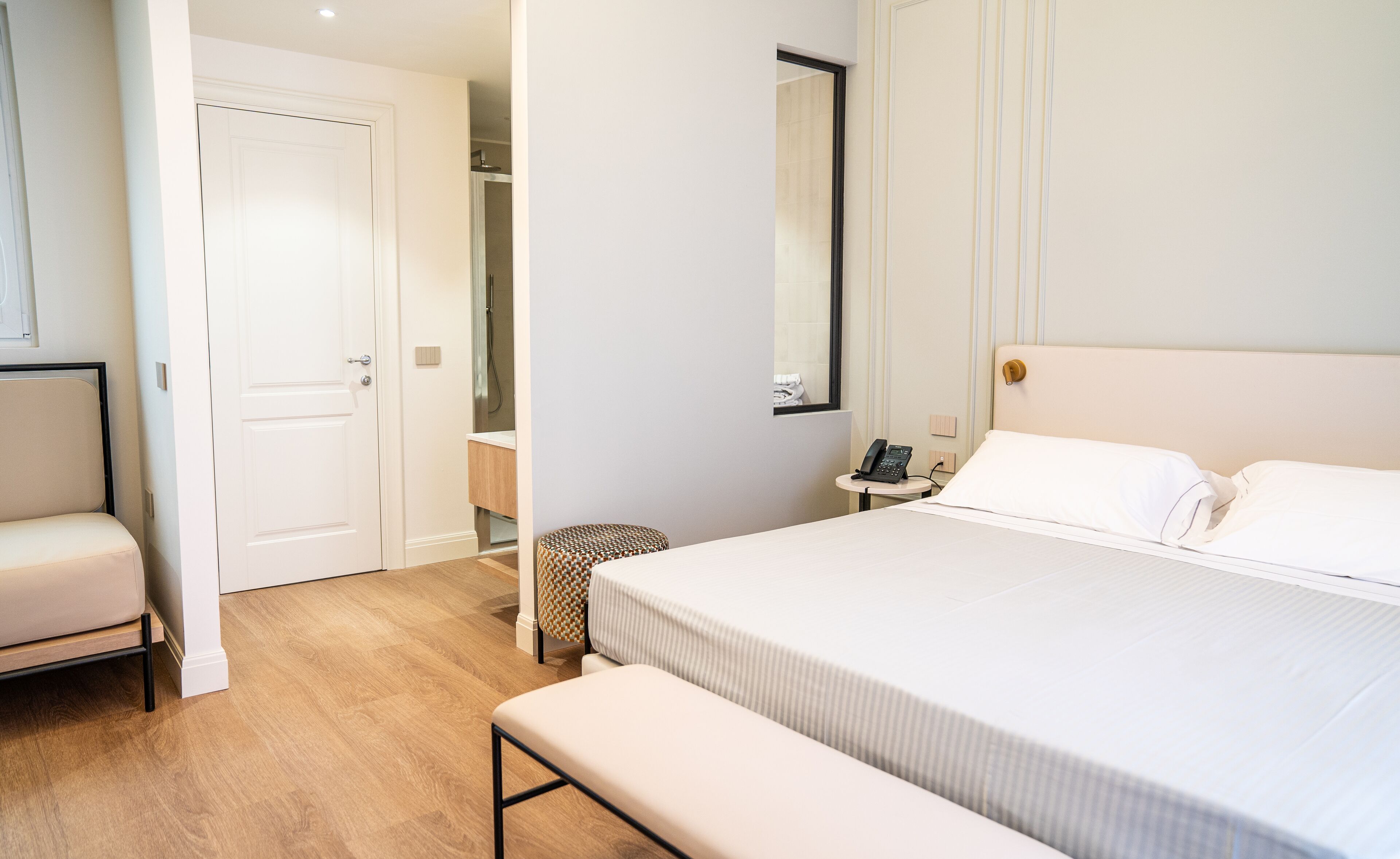 A hotel room with a bed, a bench, a bedside table with a phone, and a bathroom visible through an open door.