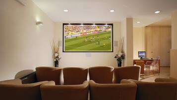 A living room with a large TV screen displaying a sports event, comfortable seating, a small desk with a computer, and a wooden door in the background.