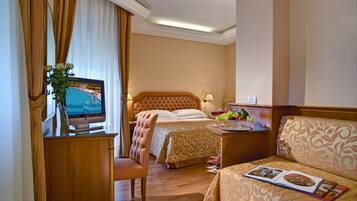 A hotel room with a bed, a desk with a television, a chair, a sofa, and a dining table with fruits.