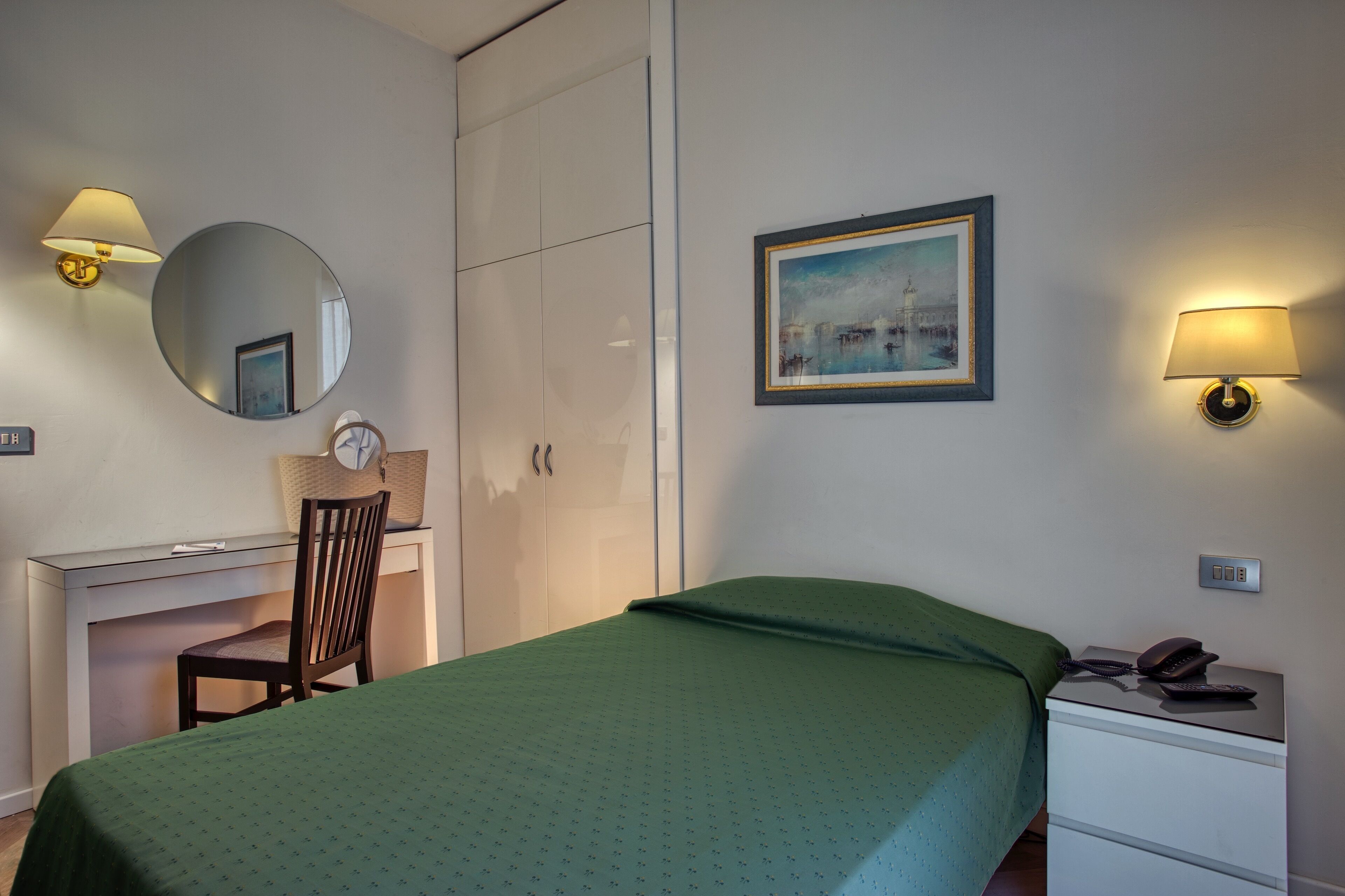 A hotel room with a bed, a desk with a chair, a wardrobe, a bedside table with a phone, and a wall-mounted lamp.