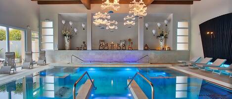 An indoor swimming pool with clear water, wooden ceiling beams, and modern lighting fixtures.
