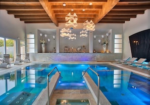 An indoor swimming pool with clear water, wooden ceiling beams, and modern lighting fixtures.