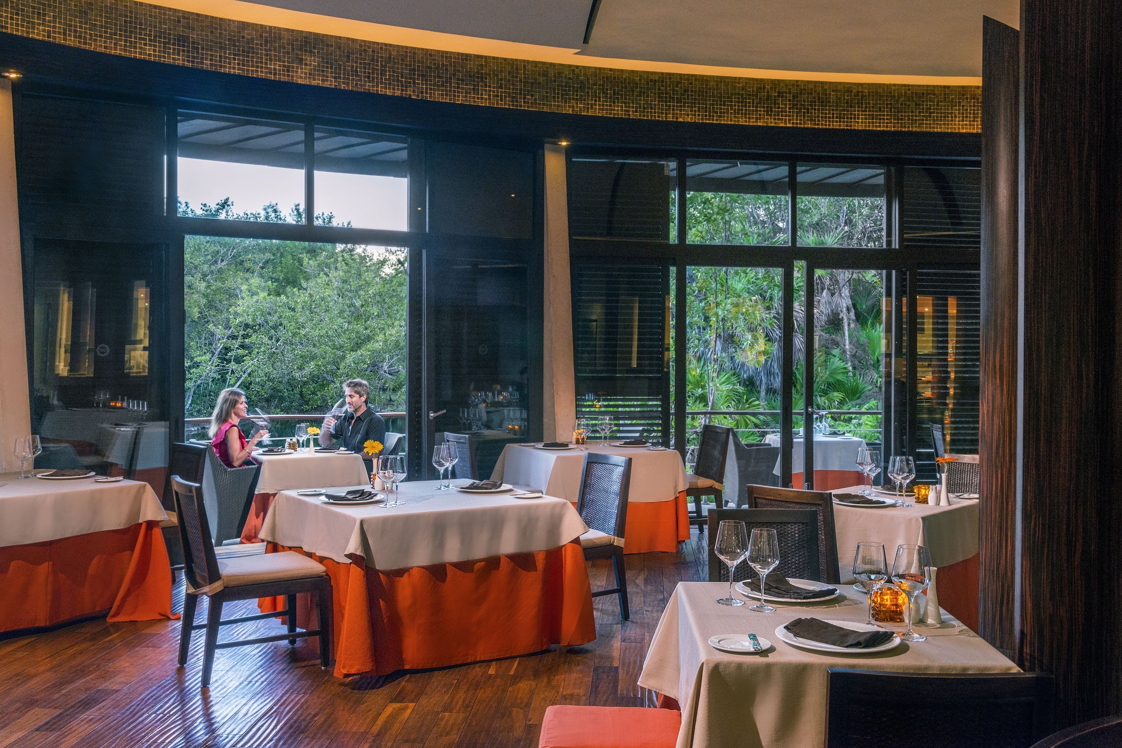 A restaurant with tables set for dining, large windows, and a view of greenery outside.