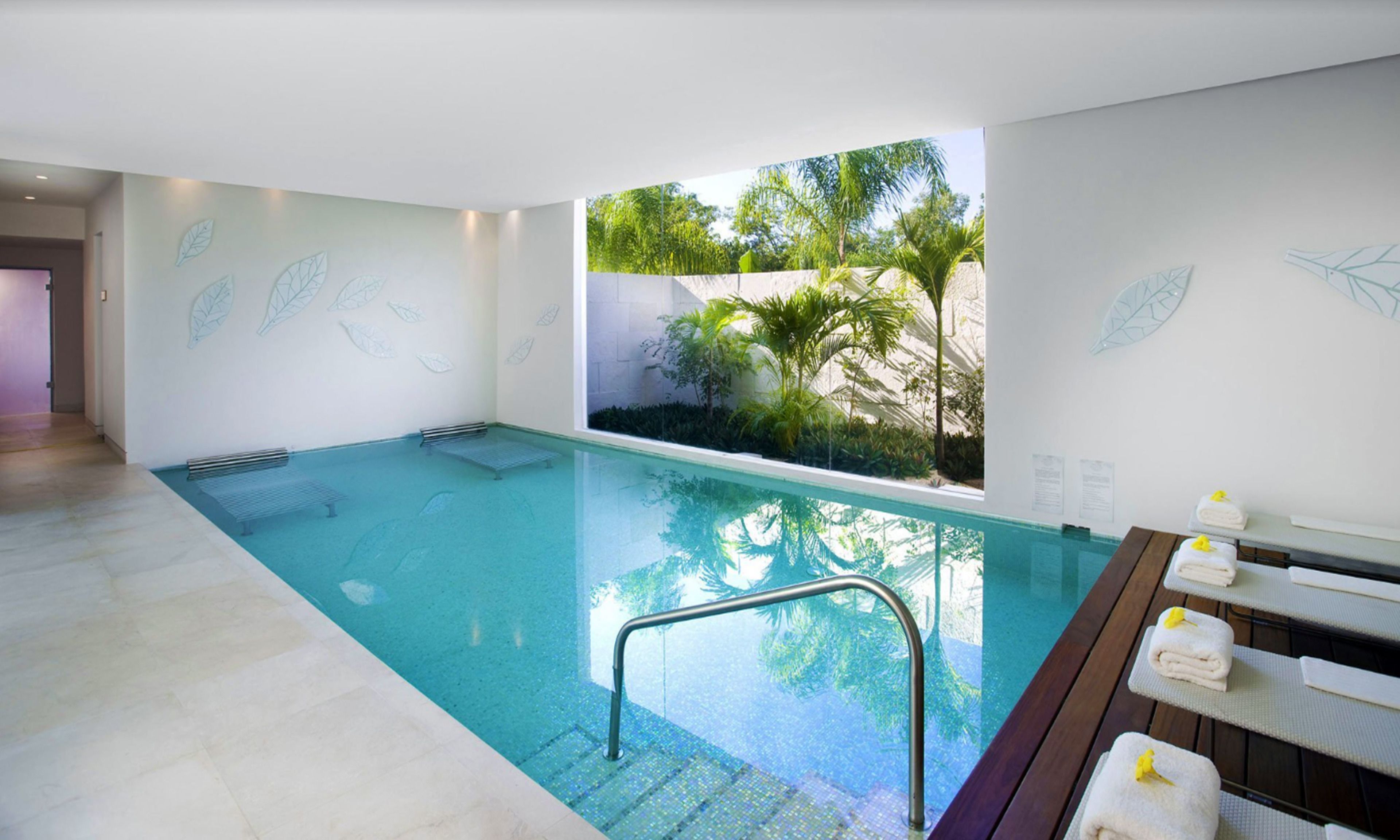 A swimming pool with a view of tropical plants, a wooden bench with towels, and a tiled floor.