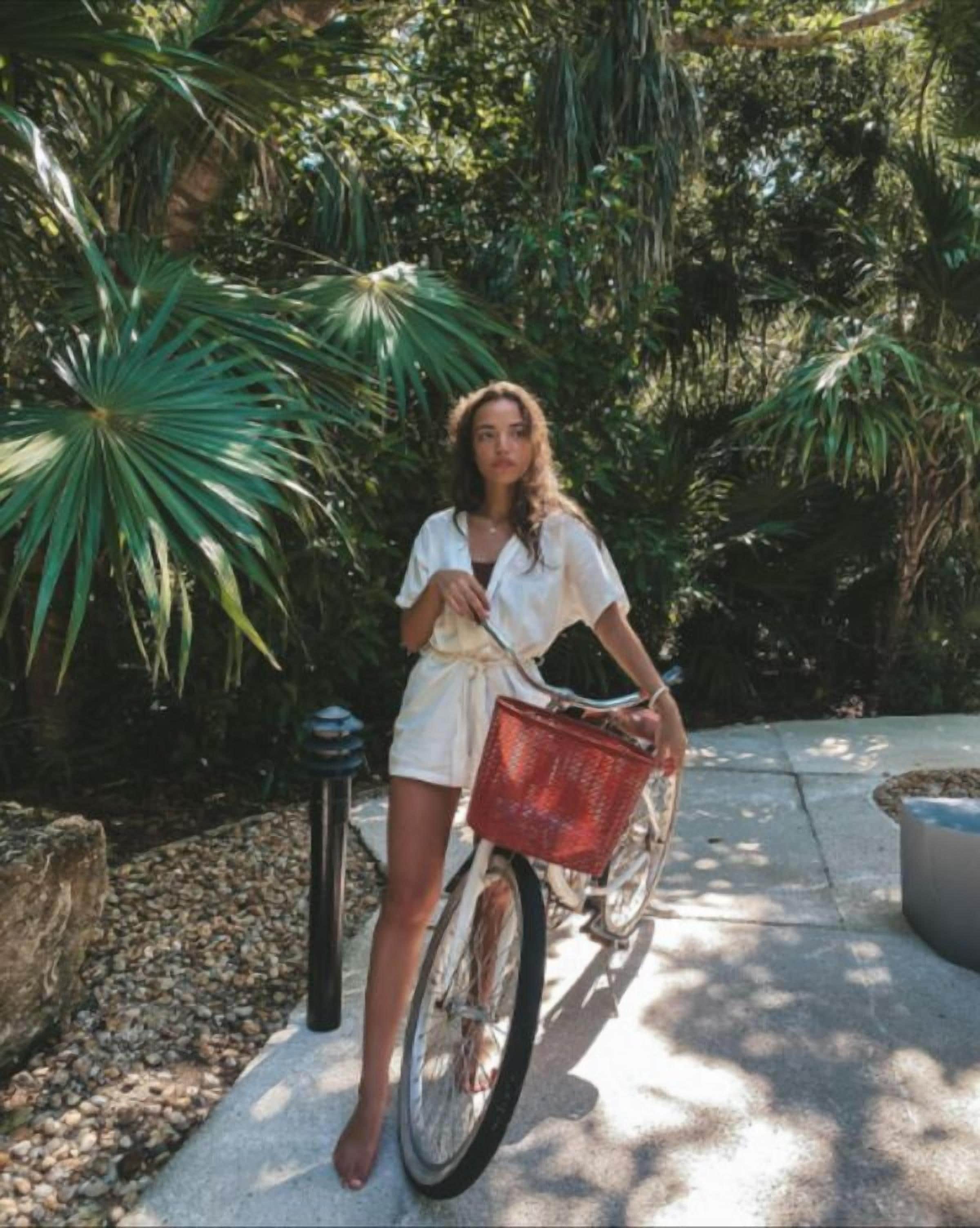 A person standing next to a bicycle with a basket, in a garden with greenery and a concrete path.
