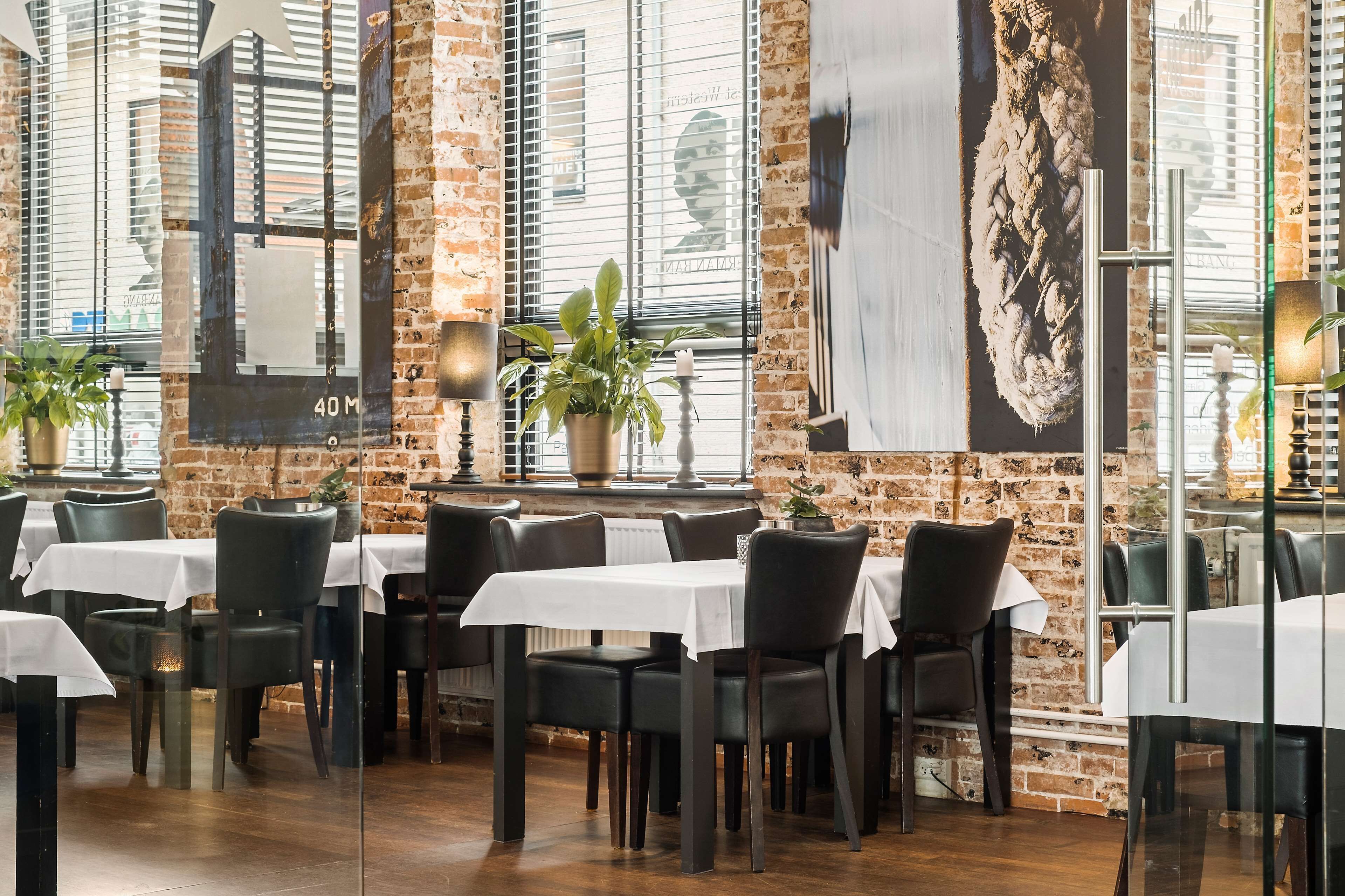 Een restaurant met bakstenen muren, grote ramen en tafels gedekt met witte tafelkleden en zwarte stoelen.