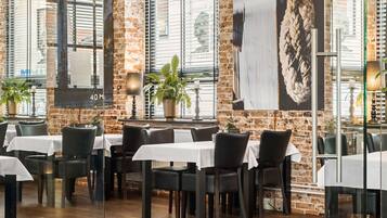 Een restaurant met bakstenen muren, grote ramen en tafels gedekt met witte tafelkleden en zwarte stoelen.