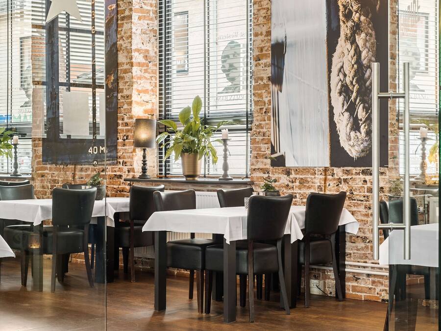 Een restaurant met bakstenen muren, grote ramen en tafels gedekt met witte tafelkleden en zwarte stoelen.