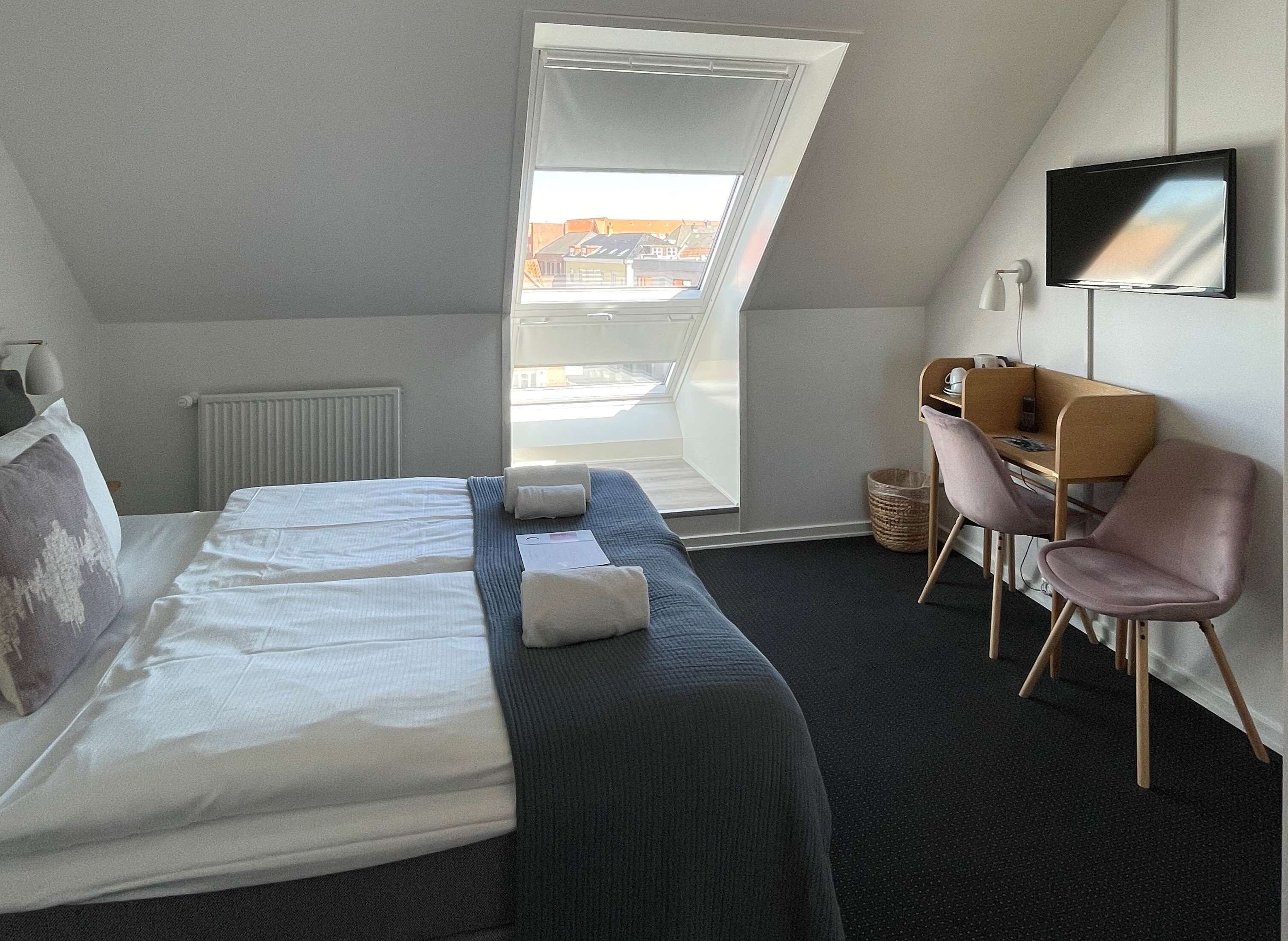 Habitación de hotel con cama, escritorio, silla, televisor y ventana con vistas a edificios.