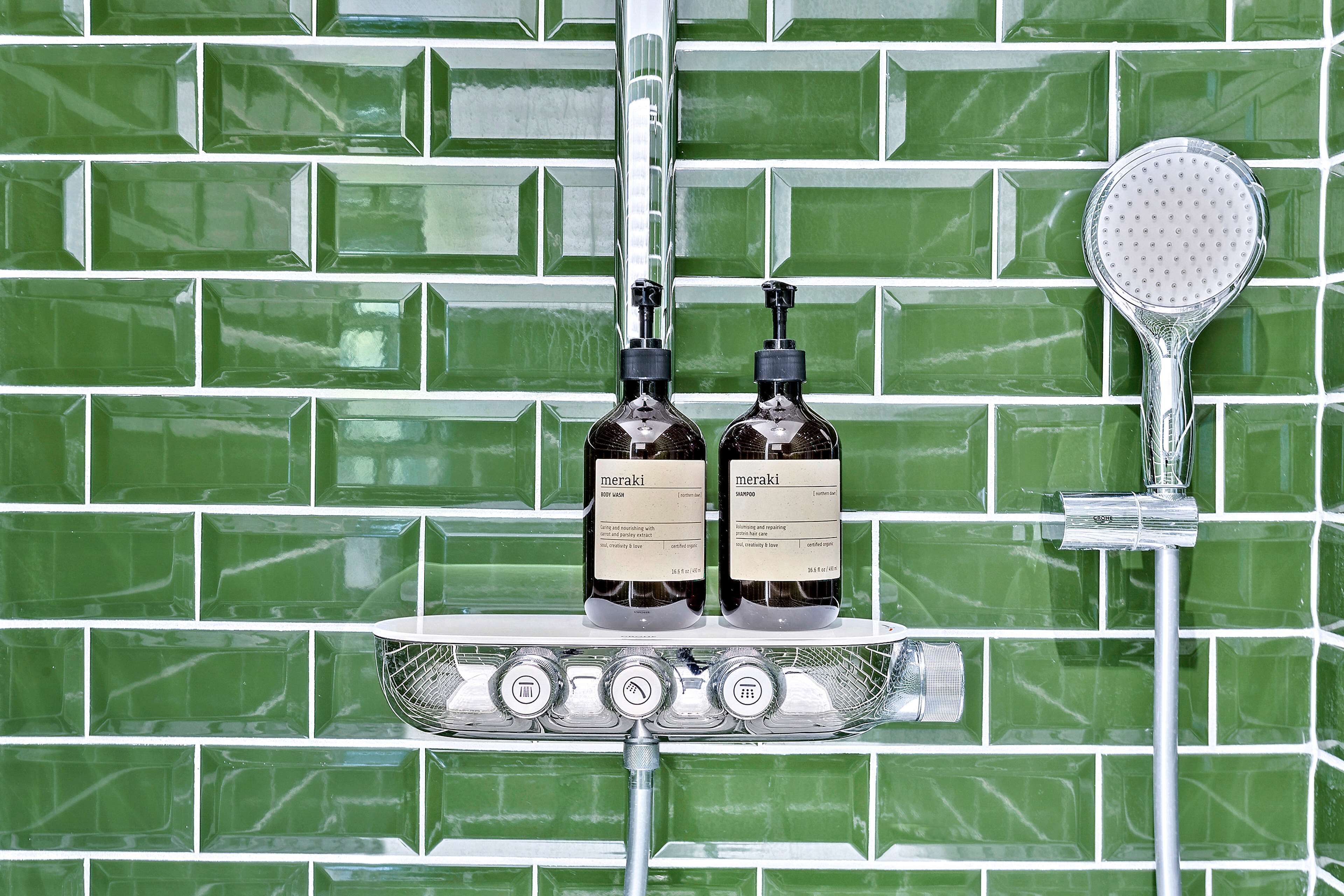 Une salle de bain avec des carreaux verts, deux bouteilles de shampoing Meraki, une pomme de douche et un pommeau de douche à main.