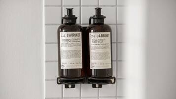 Two bottles of L'A BRUKET shampoo and body wash mounted on a tiled wall.