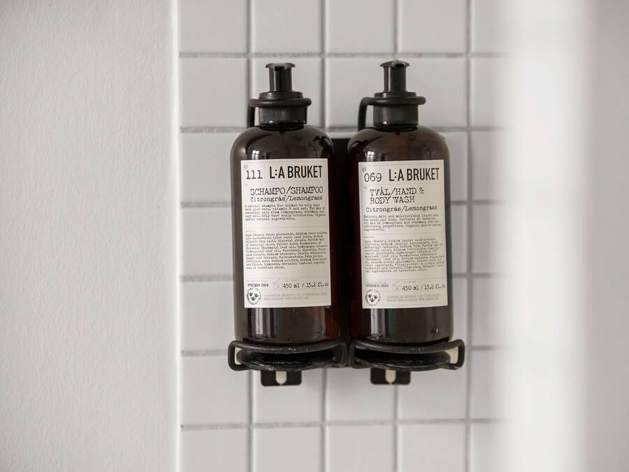 Two bottles of L'A BRUKET shampoo and body wash mounted on a tiled wall.