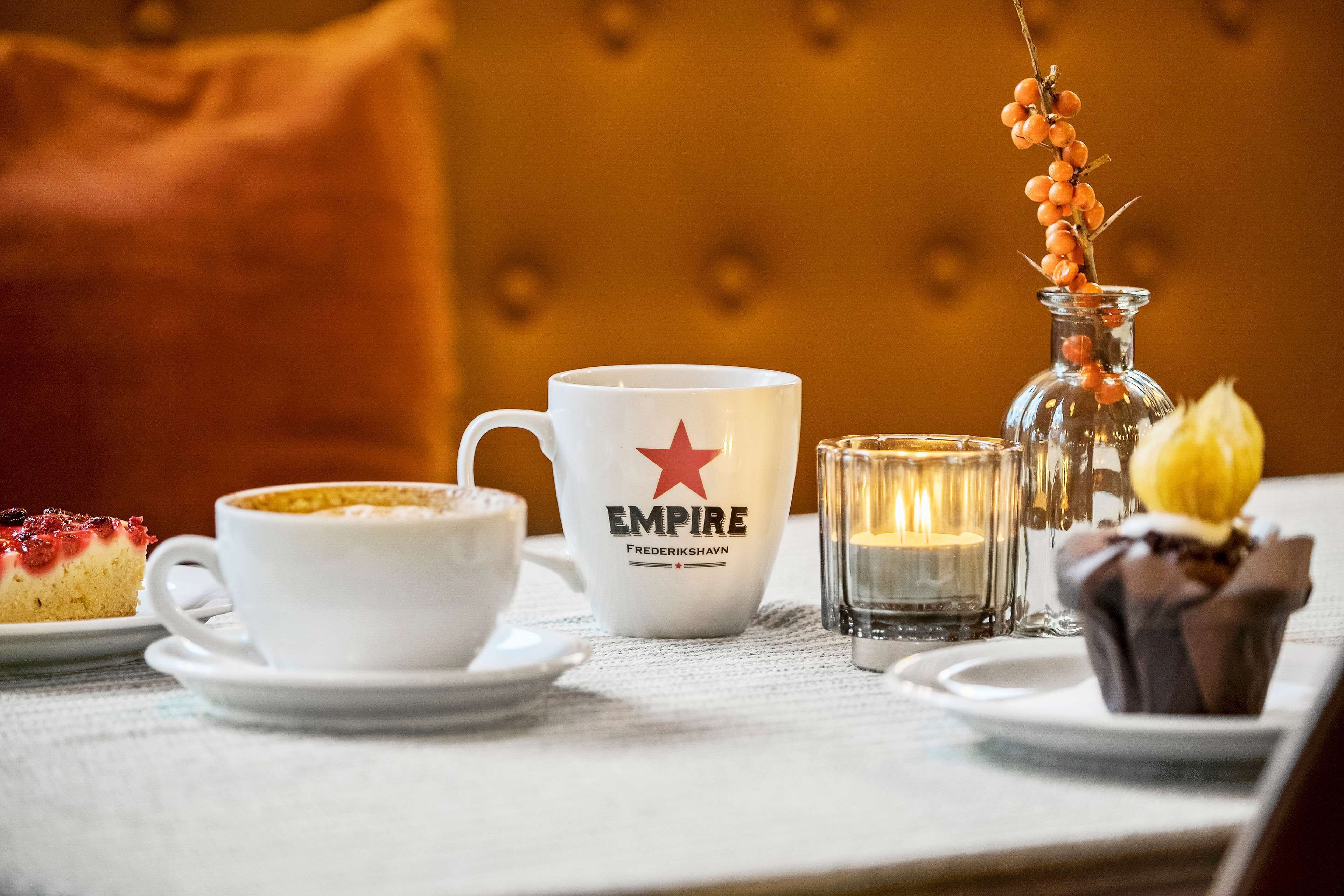 Una mesa con una taza blanca que dice "EMPIRE", un candelabro de cristal y un plato de postre con una porción de pastel.