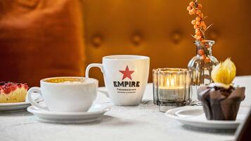 Una mesa con una taza blanca que dice "EMPIRE", un candelabro de cristal y un plato de postre con una porción de pastel.
