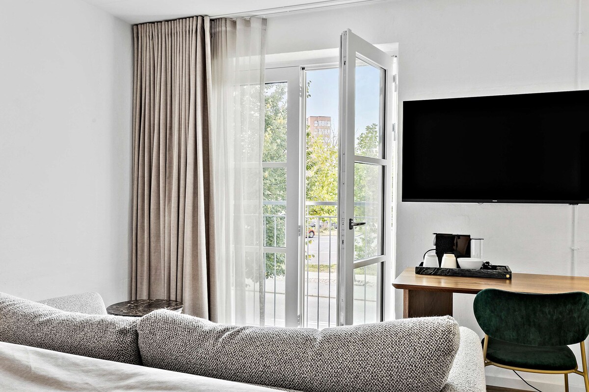 A modern hotel room with a large window, a flat-screen TV, a wooden desk, and a green chair.