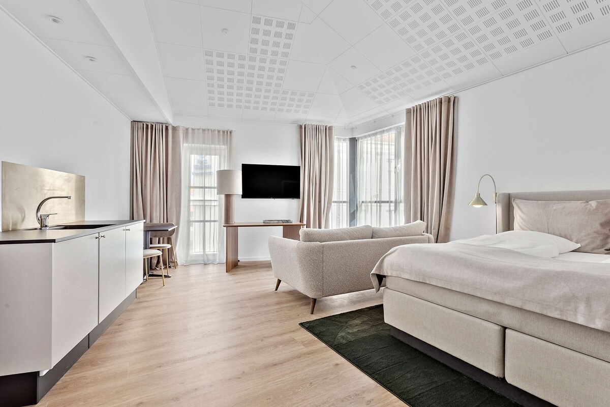 A modern hotel room with a large bed, a flat-screen TV, a desk with a chair, and a kitchenette.