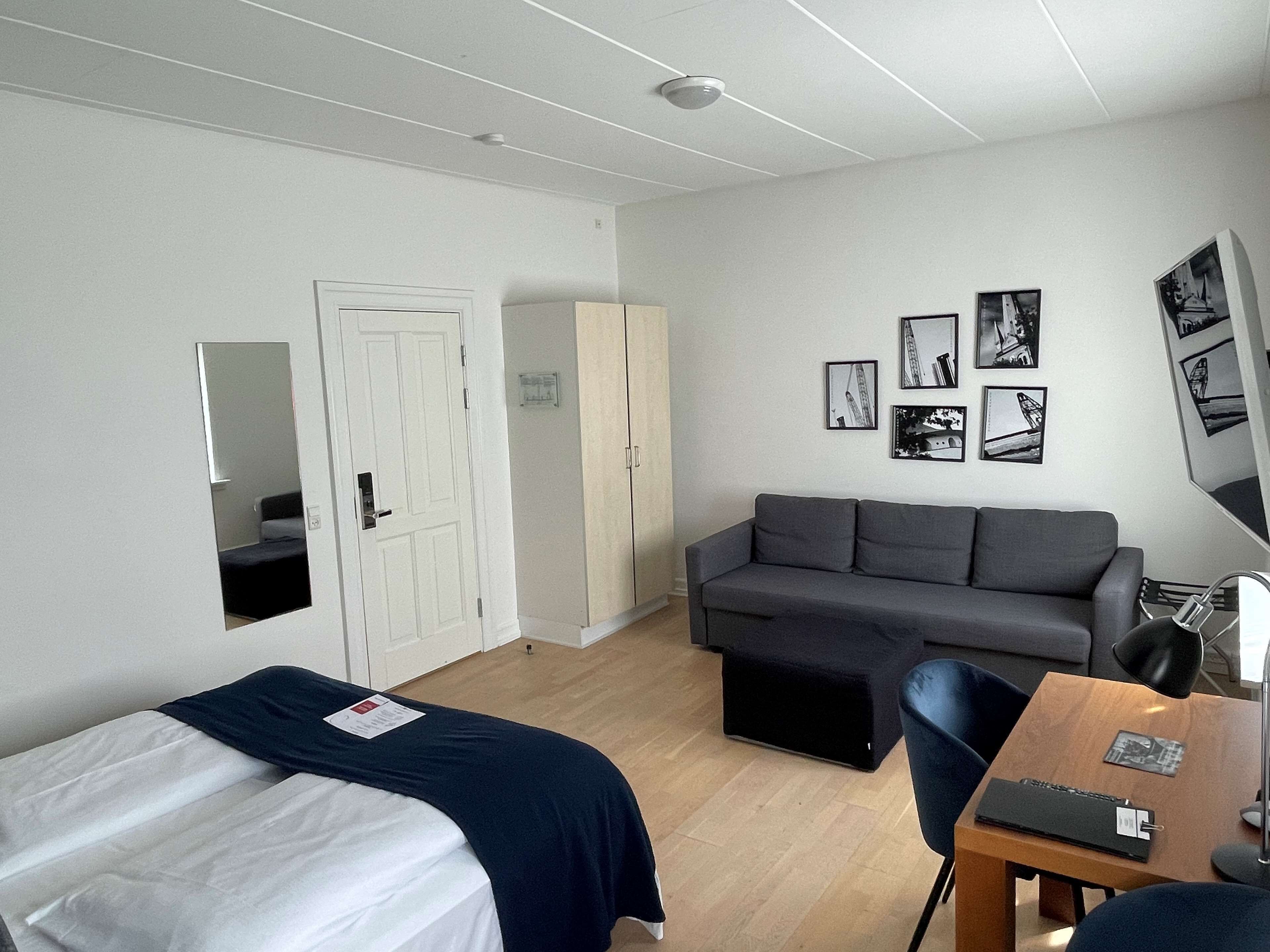 Una habitación con dos camas individuales, un sofá gris, una mesa de madera con una lámpara y un espejo en la pared.