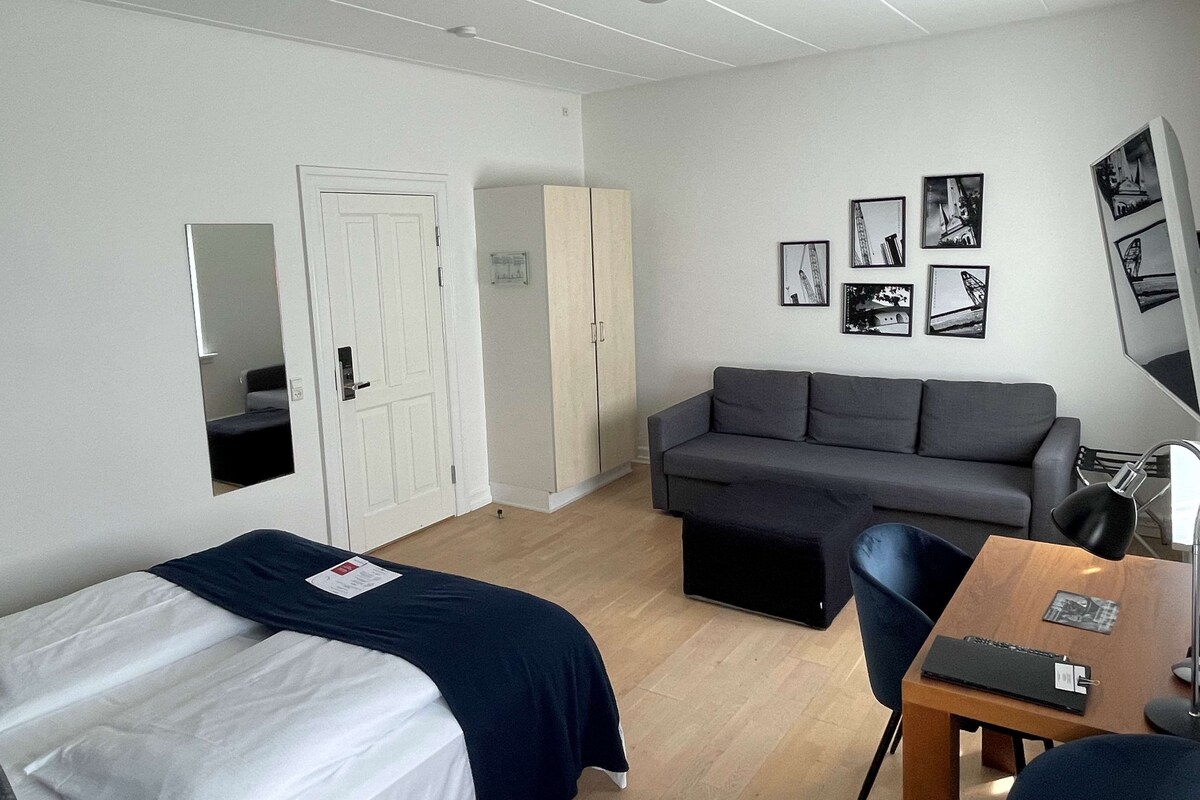 A room with two single beds, a grey sofa, a wooden table with a lamp, and a mirror on the wall.