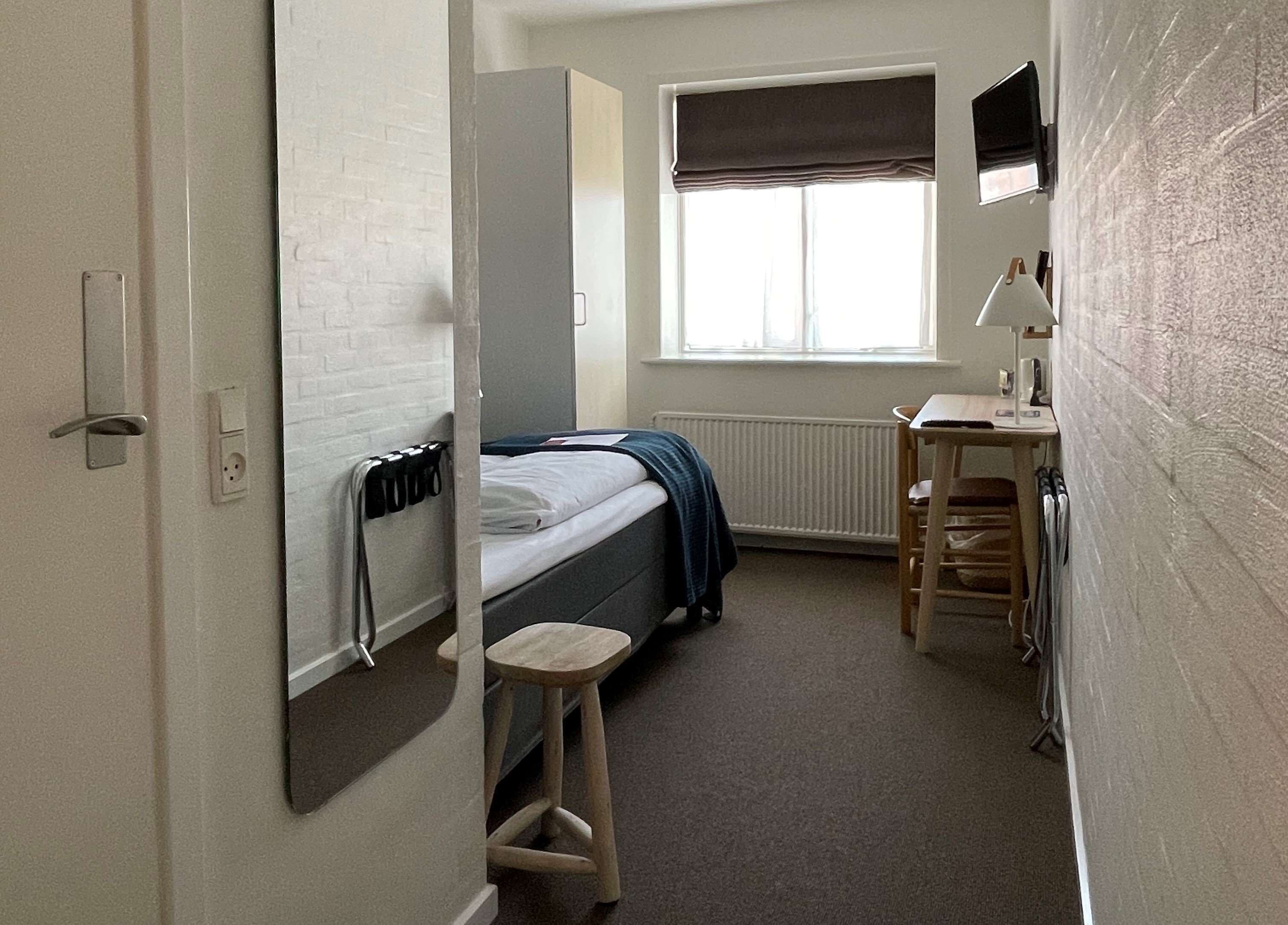 A compact room with a bed, a desk, a chair, a radiator, and a window with a blind.