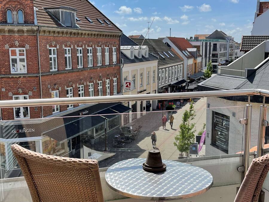 A balcony with wicker chairs and a table, overlooking a street with buildings and shops.