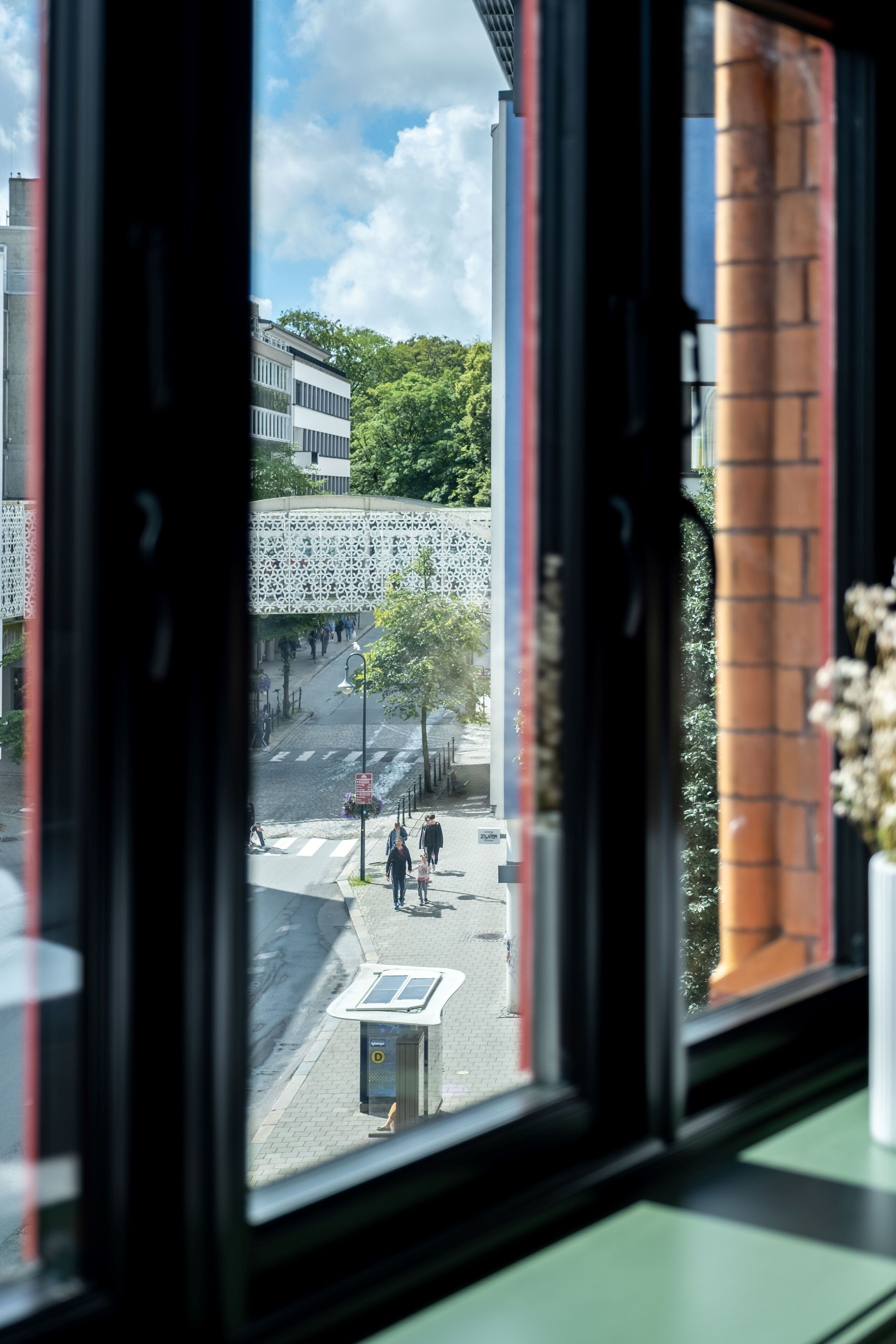 Udsigt fra et vindue over en bygade med fodgængere, en busholdeplads og en bygning med en usædvanlig facade.