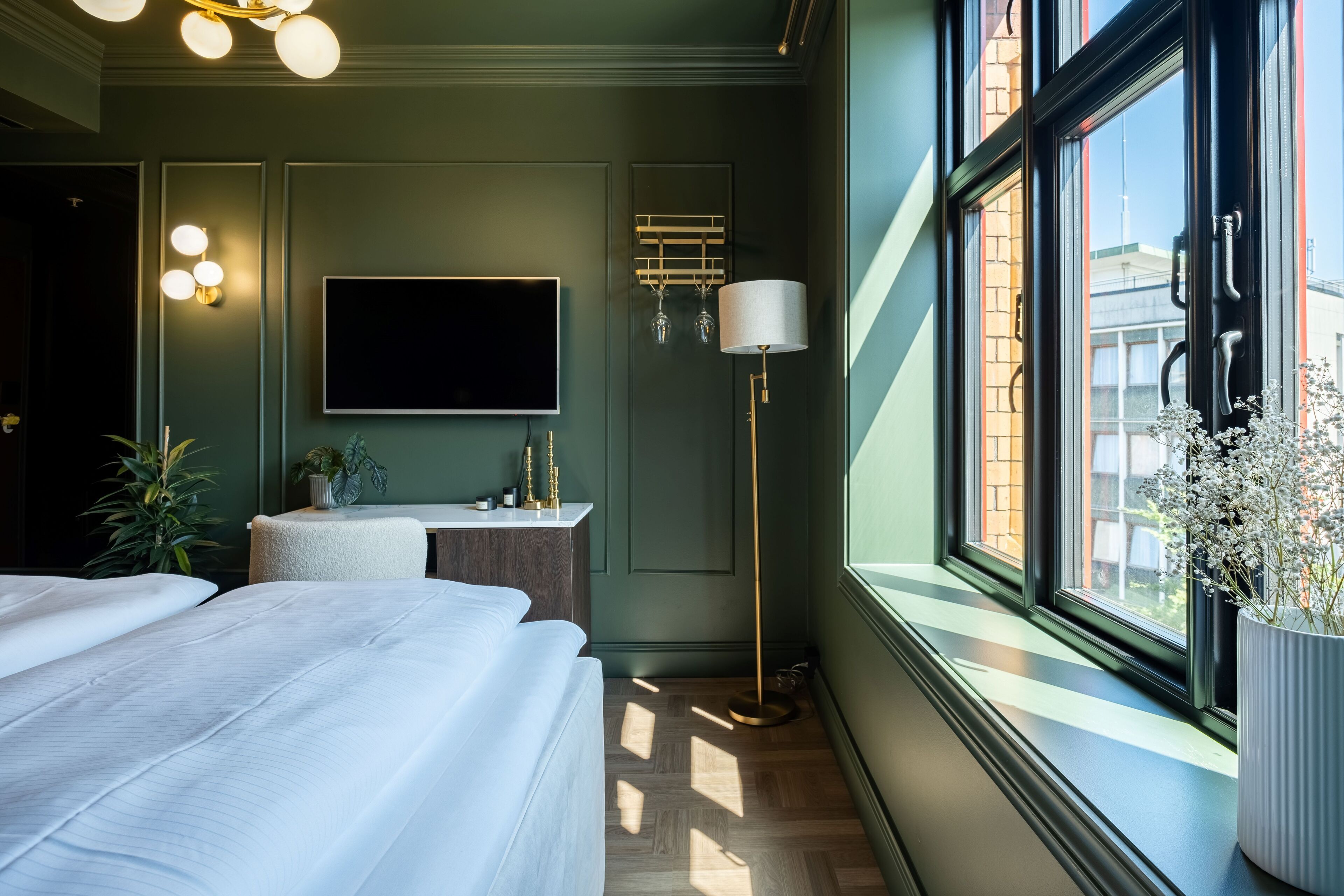 A modern hotel room with a large bed, a flat-screen TV, a bedside table, a floor lamp, and a view of the city through the window.