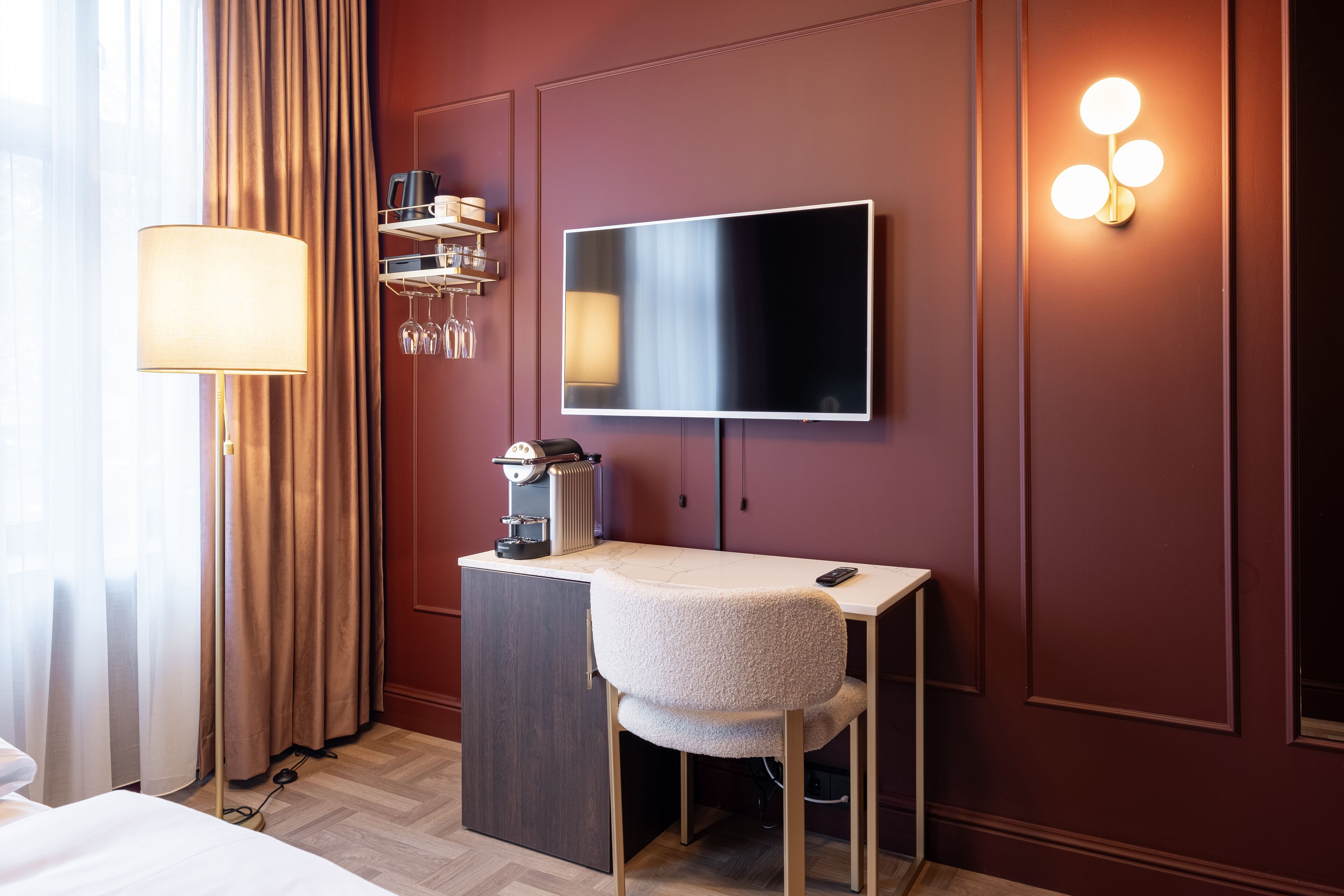 Camera d'albergo con un letto, una scrivania con una sedia, una TV a schermo piatto, una macchina per il caffè, una lampada e uno scaffale a parete con bicchieri da vino.