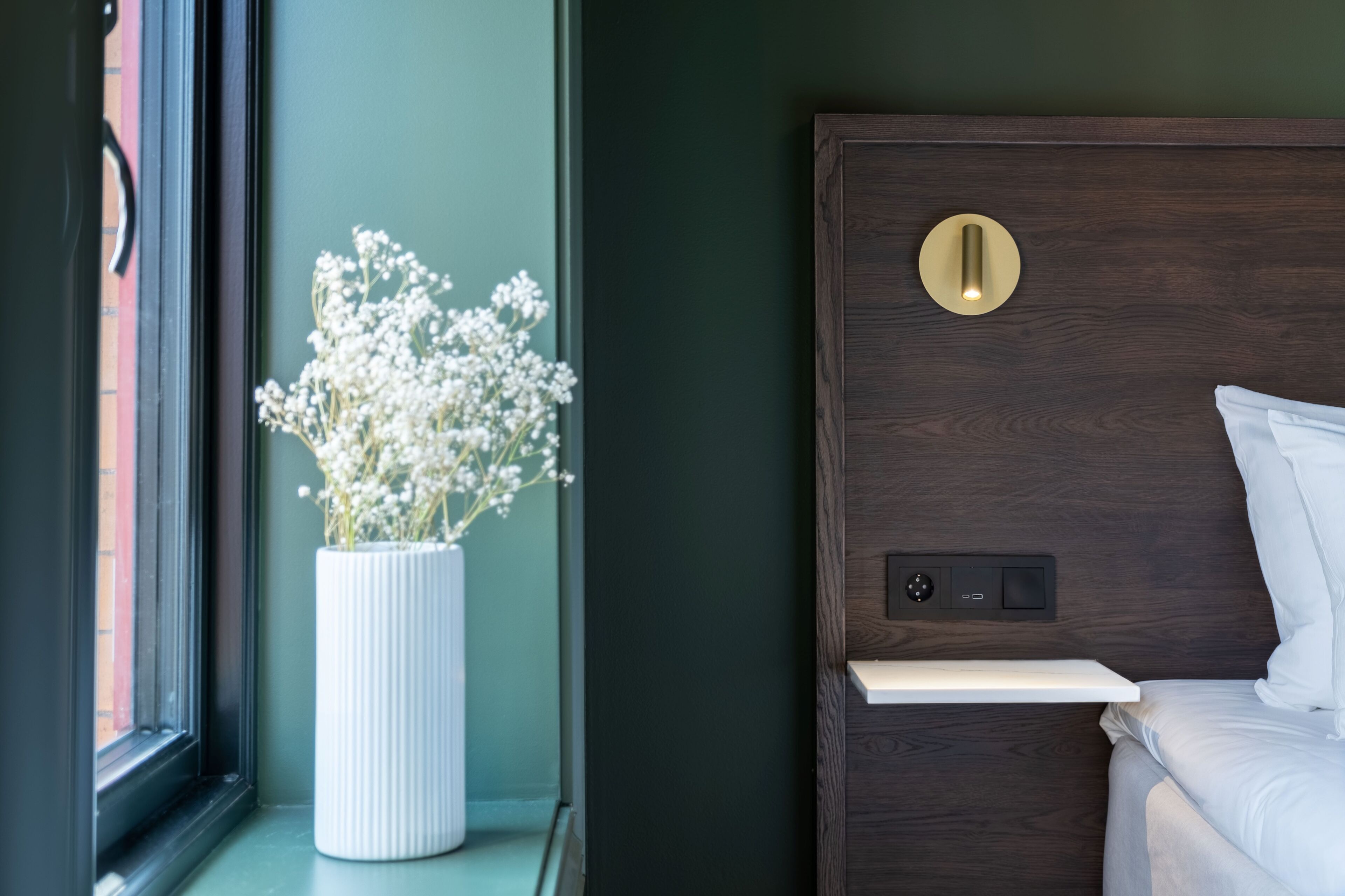 A bedroom with a dark wooden headboard, a bedside table, and a white vase with flowers.