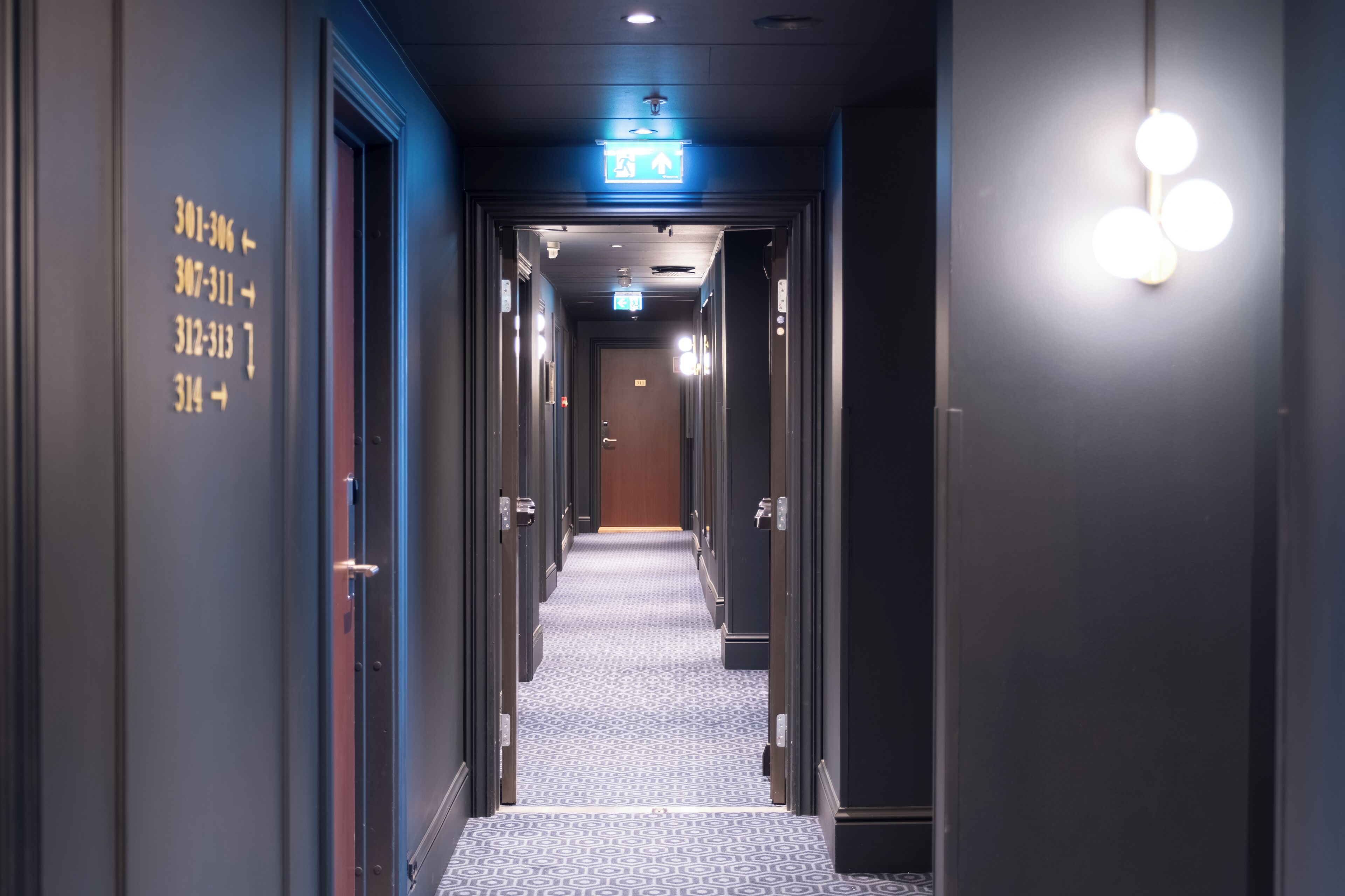 A hotel hallway with numbered doors, an exit sign, and a patterned carpet.