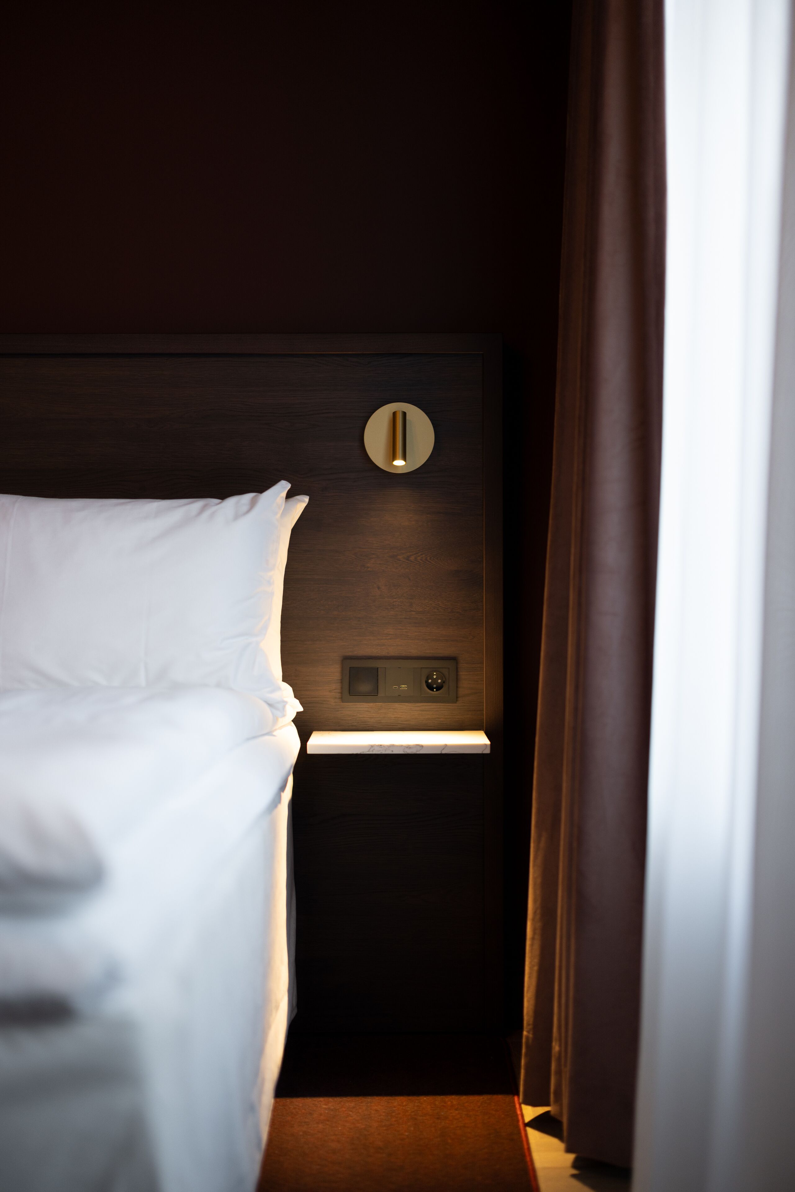 A hotel room with a neatly made bed, a bedside table with a lamp, and a wall-mounted control panel.