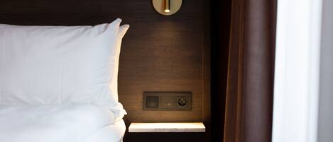 A hotel room with a neatly made bed, a bedside table with a lamp, and a wall-mounted control panel.