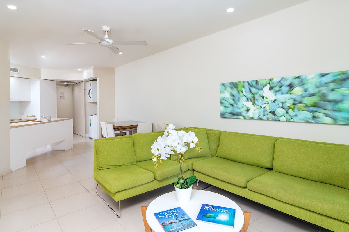 A modern living room with a green sofa, a white coffee table with books, and a ceiling fan.
