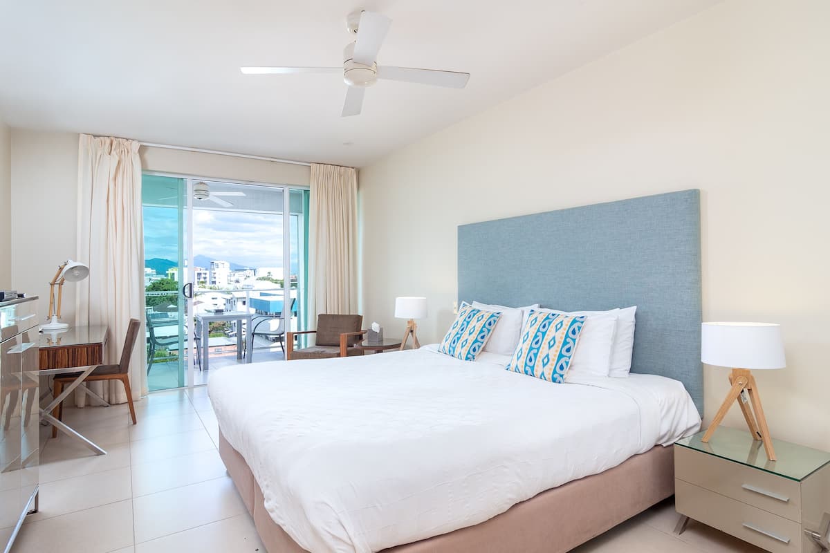 A hotel room with a large bed, a desk, a chair, a glass table, and a ceiling fan.