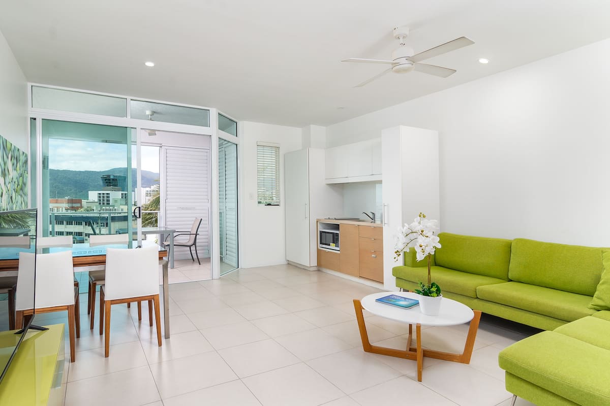 A modern living room with a green sofa, a dining area with white chairs, a kitchen with white cabinets, and a view of the mountains through large windows.