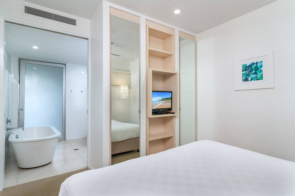 A modern hotel room with a large bed, a television on a wooden shelf, a bathroom with a bathtub, and a framed picture on the wall.
