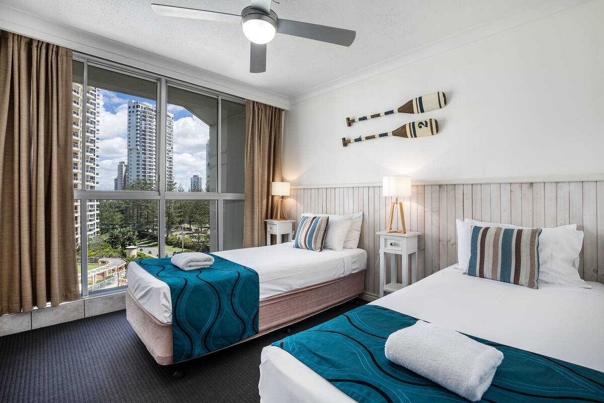 A hotel room with two beds, a large window with curtains, a ceiling fan, and a view of buildings outside.