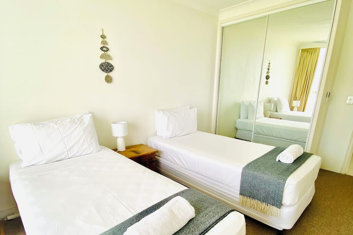 A hotel room with two beds, a ceiling fan, a bedside table with a lamp, and a mirror reflecting the room.